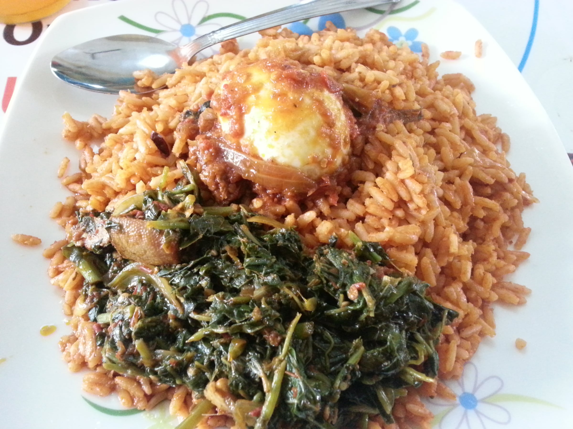 A plate of orange-red jollof rice served with greens and an egg