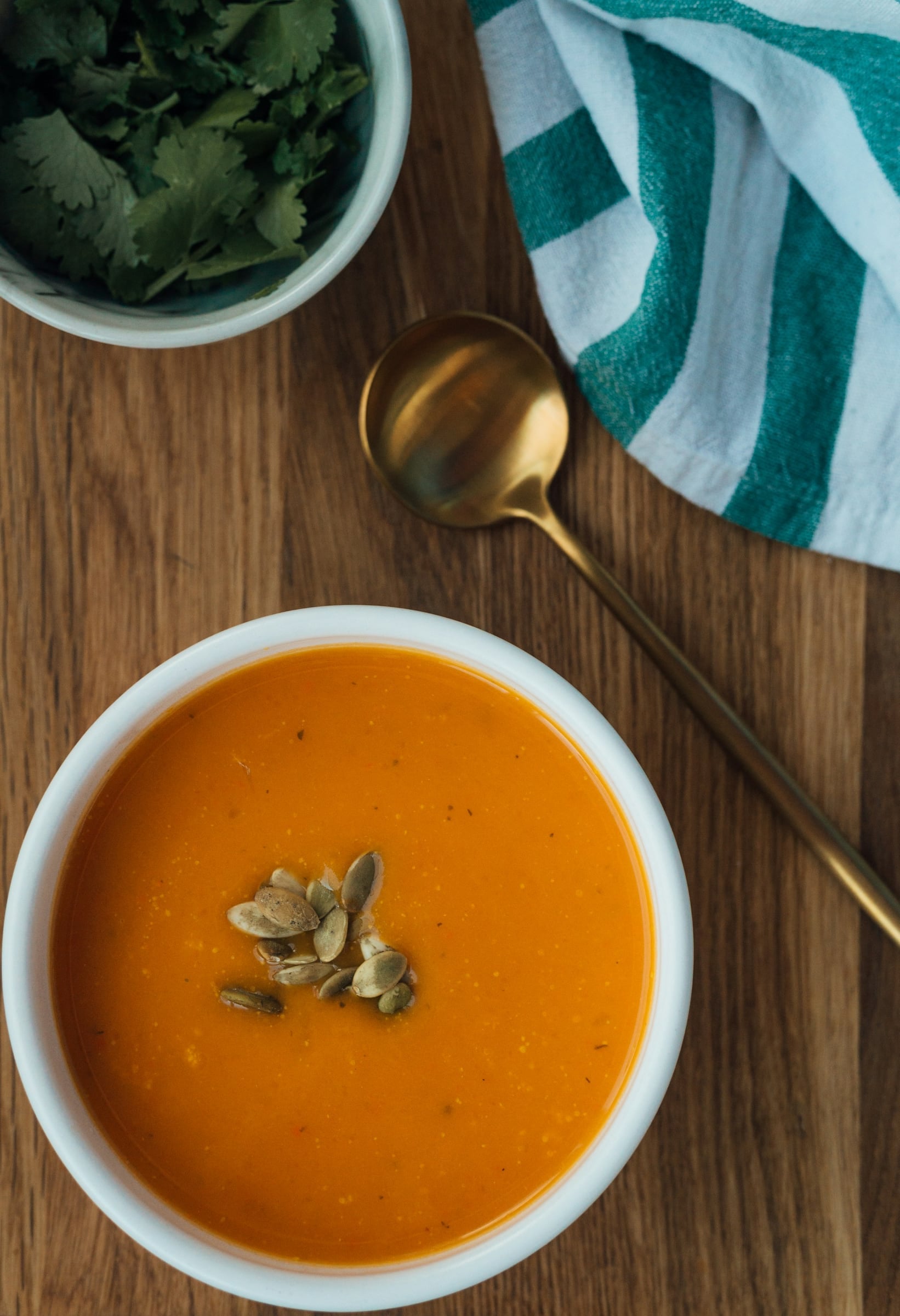 A bowl of smooth orange soup topped with toasted pumpkin seeds, with a gold spoon on a wooden table