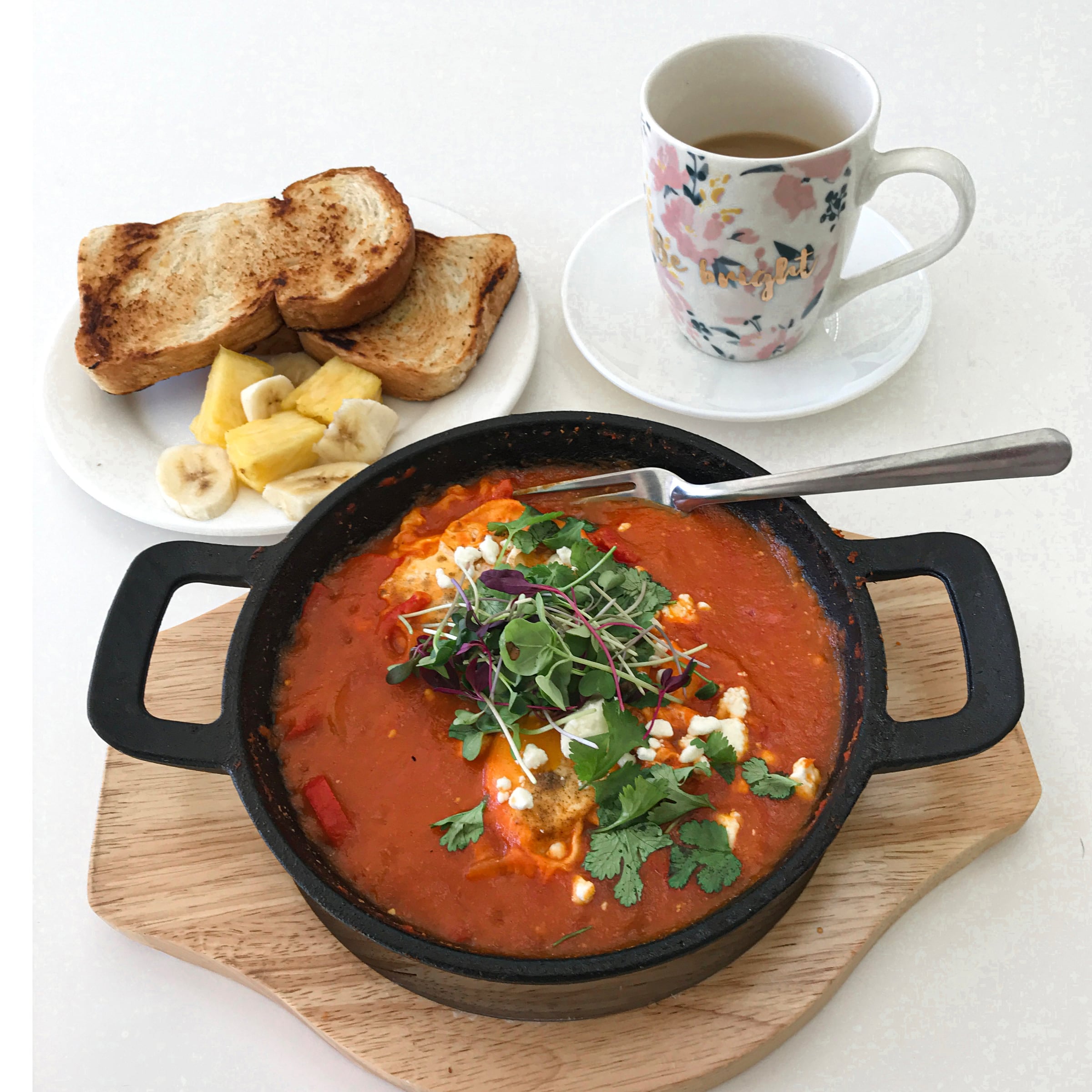 A black pan of shakshuka: eggs poached in red tomato sauce, scattered with herbs and feta, with toast alongside